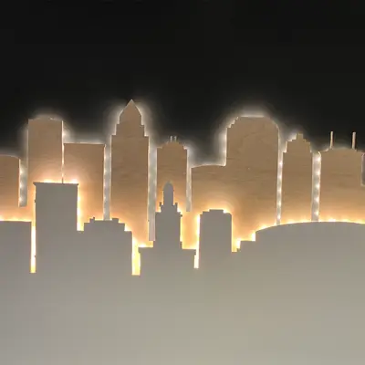 A multi-layered wooden skyline silhouette installation on a wall, backlit with warm white lighting that glows against a dark background.