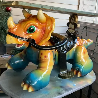 A repurposed, vintage coin-operated kids' riding toy customized into a colorful triceratops dinosaur. It has a vibrant orange and yellow head, a blue and green body with spots, large green eyes, a black saddle, and a small painted character cutout on the back.