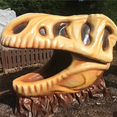 A large, realistic sculpture of a Tyrannosaurus Rex dinosaur skull, detailed with weathered bone textures and shades of tan and brown, sitting outdoors on a mulched surface.