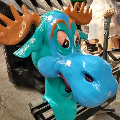 A large, brightly painted paper-mache or sculpted prop head of a cartoon moose in a workshop. It is turquoise blue with orange antlers, and has a playful, slightly weary expression with large eyes. It is mounted on a stand, surrounded by other production materials.